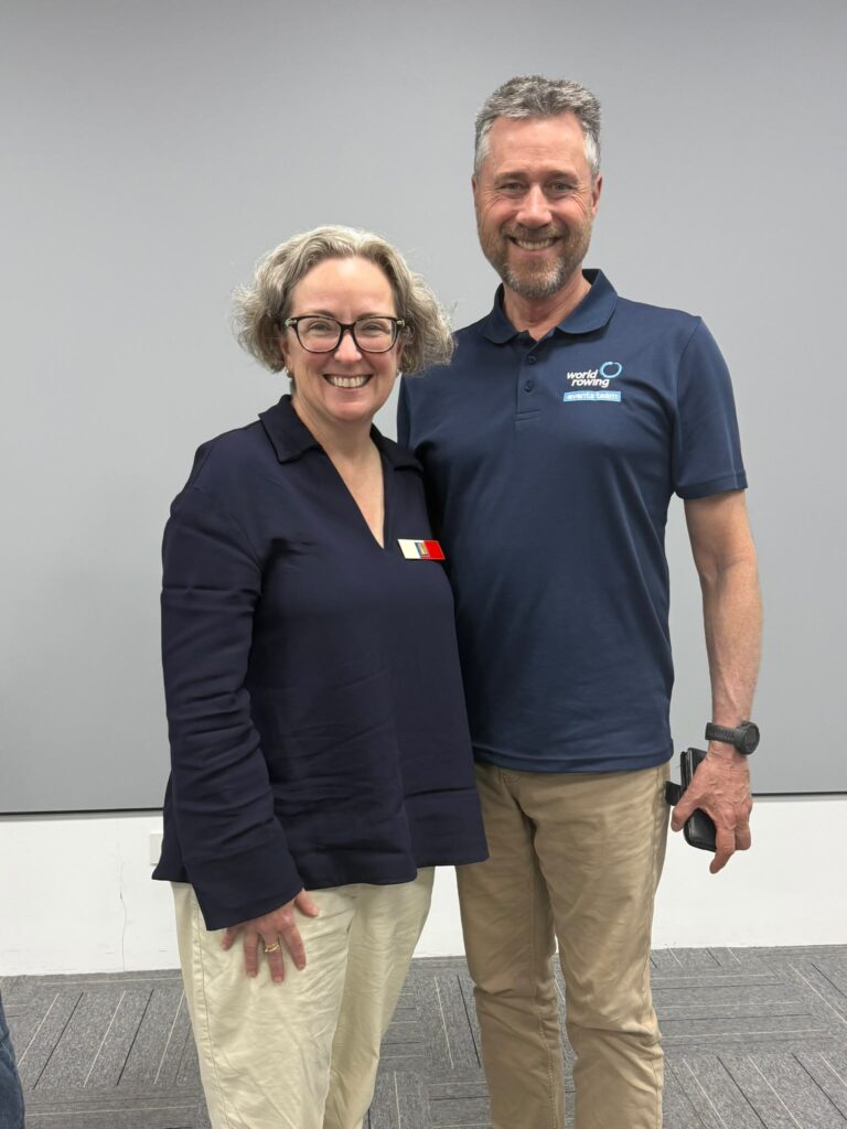 Two people smiling in a meeting room. One wears a navy top with a Rowing SA badge, and the other wears a navy World Rowing Events Team polo.
